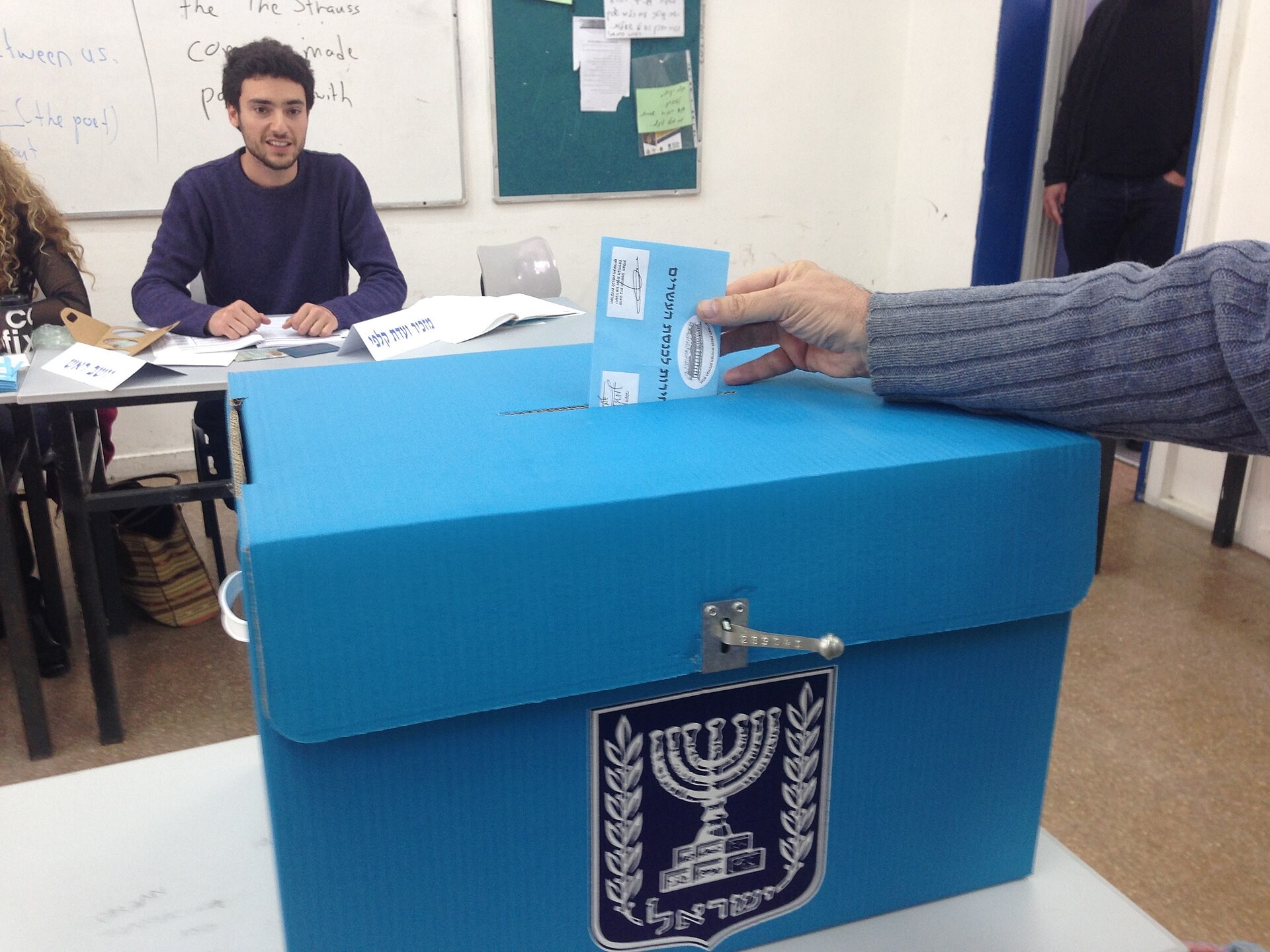 En hand håller ett blått kuvert över en blå valurna med Israels statsvapen.