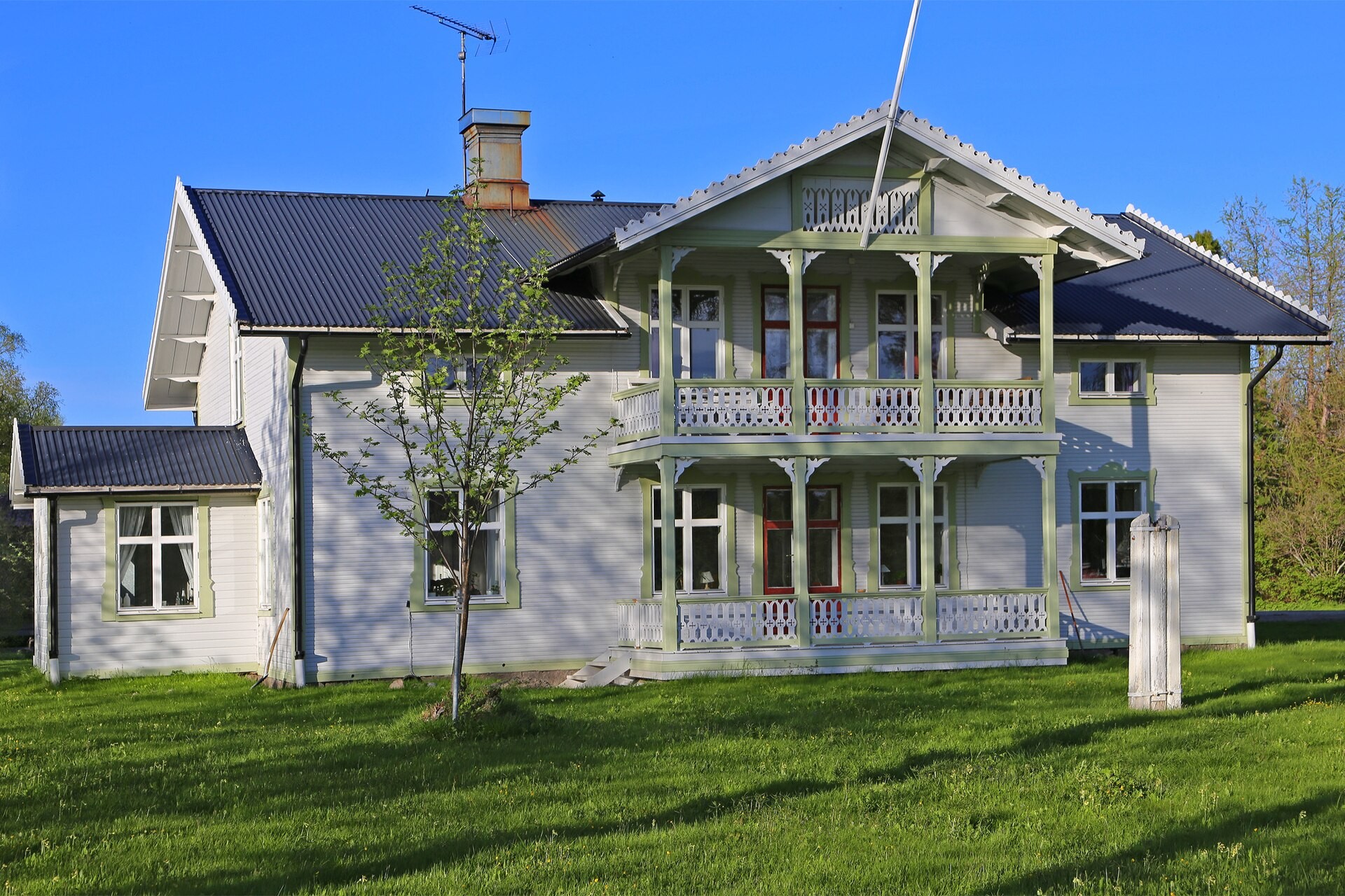 Ljust trähus med gröna detaljer, veranda och balkong framför en gräsmatta.