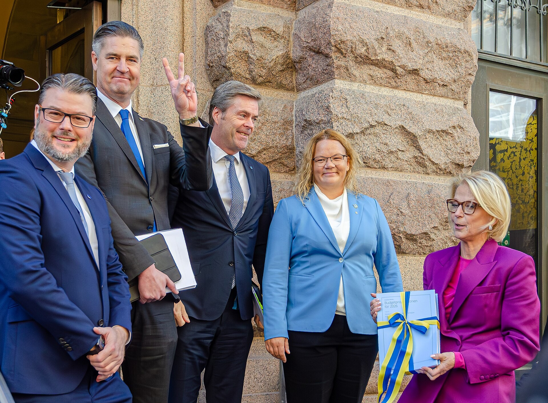 Fem personer står framför en stenfasad. En av dem håller en budgetproposition med blågula band.