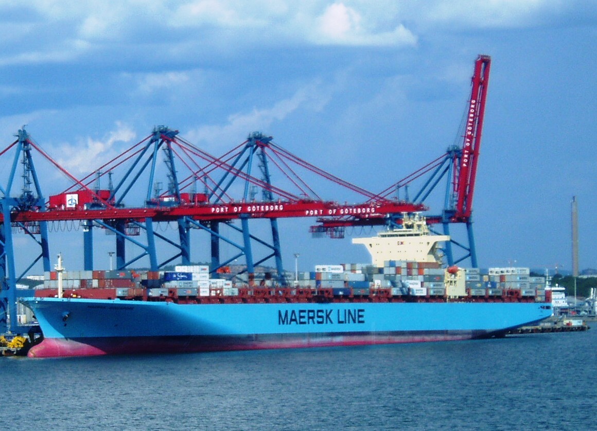 Ett blått containerfartyg vid kaj under stora röda och blå hamnkranar.